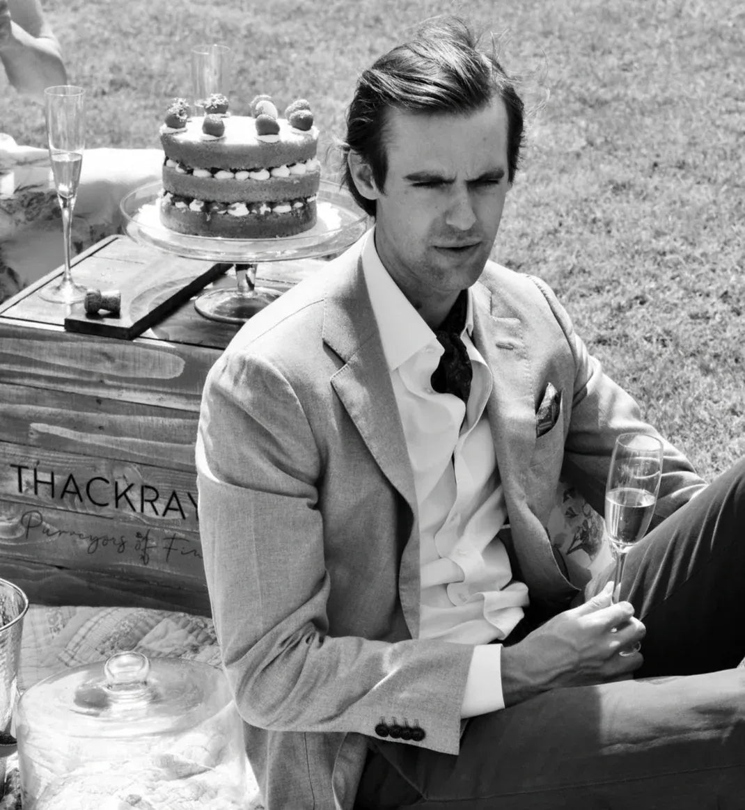 Man in formal attire sitting outdoors with a cake and champagne glass

